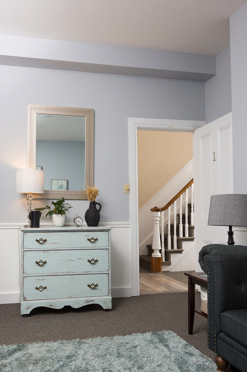 Cozy therapy room featuring a light blue dresser with decorative items, a mirror, and a plant, with a staircase visible in the background, reflecting a welcoming environment for mental health services.