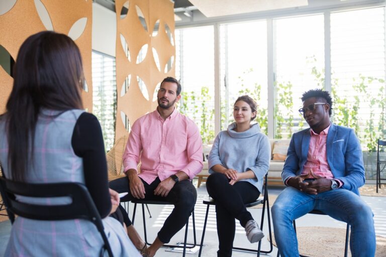 Group therapy session with four individuals seated in a bright, modern space, discussing mental health and mindfulness techniques.