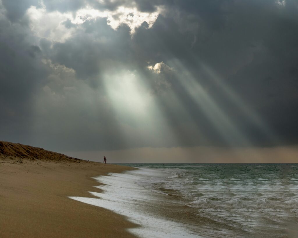 Beach scene with soft waves lapping at the shore, a lone figure walking along the sand under dramatic, cloudy skies with rays of light breaking through, symbolizing hope and healing for women seeking support in the Worthy Women’s Group.