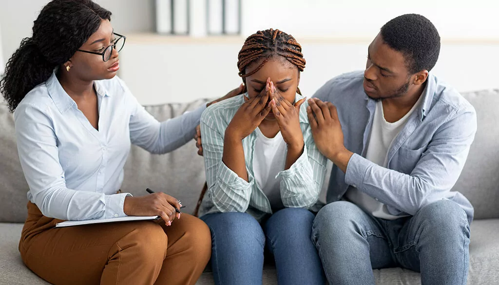Therapist providing compassionate support to a grieving individual, with a comforting presence from a supportive friend during a counseling session.
