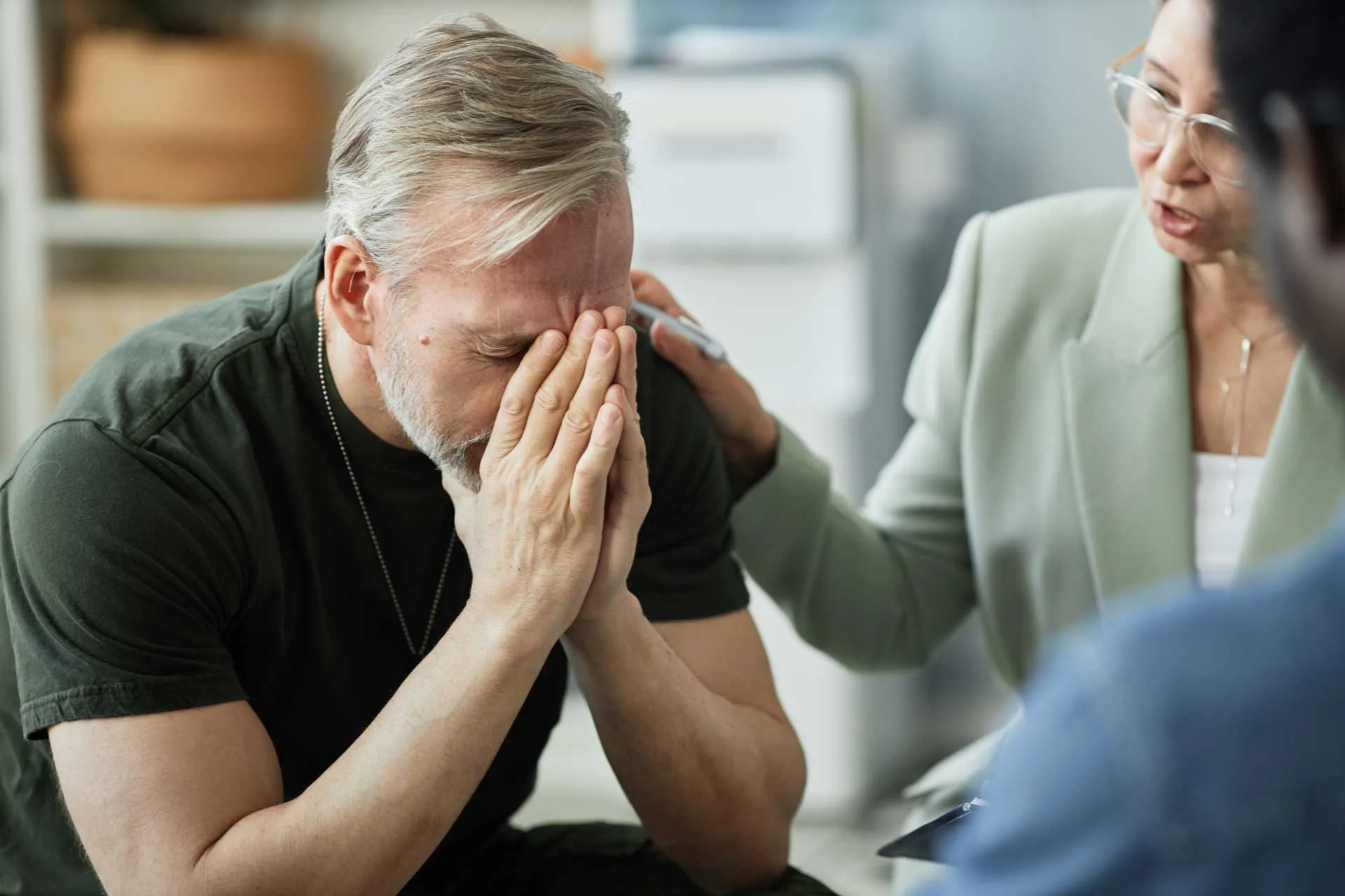 Man with gray hair and beard, sitting with head in hands, expressing distress during a therapy session, while a compassionate therapist offers support, emphasizing emotional healing and trauma-informed care.