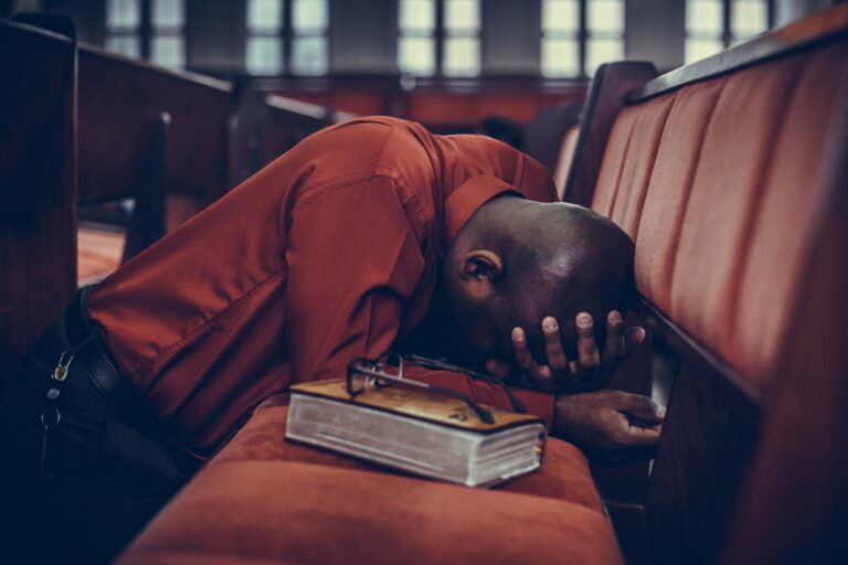 Man in red shirt sitting in church pew, head down in prayer or meditation, with a closed book beside him, reflecting themes of stress relief and emotional support in mental health.