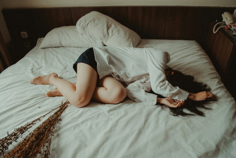 Person curled up on a bed, wearing a white shirt, conveying feelings of anxiety and distress, with dried flowers on the bed, reflecting themes of mental health and emotional well-being.