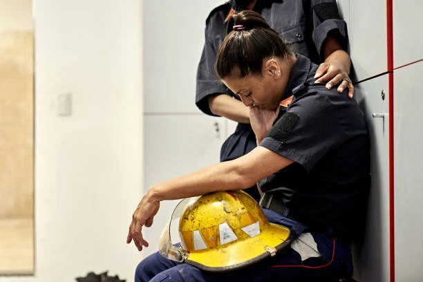 A firefighter in uniform sits on the floor with a yellow helmet resting on their lap, appearing distressed, while another person stands beside them with a comforting hand on their shoulder in an indoor setting.