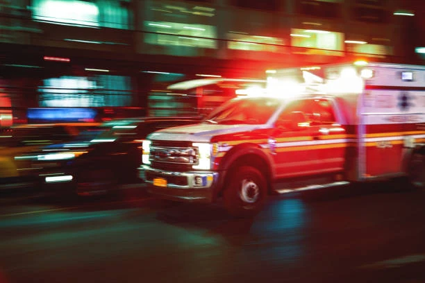 A red and white ambulance speeds through a city street at night with flashing emergency lights, creating motion blur against illuminated buildings and passing traffic in the background.